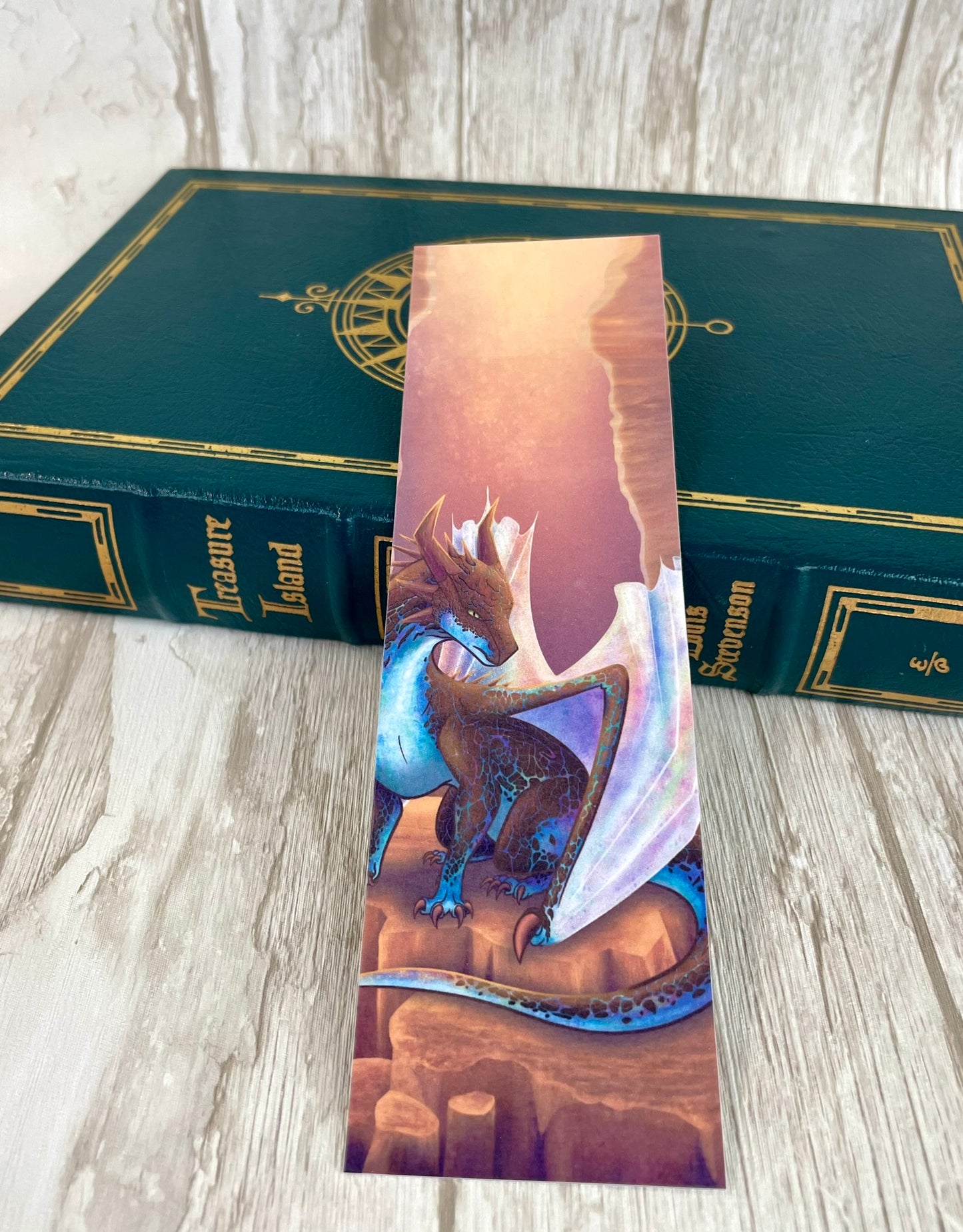A 2 by 7 inch bookmark featuring a sitting, side profile of a dragon that has the features of a boulder opal with hues of blue, green, purple, and pink on the underbelly and cracks of the rough, brown hue, rock scales. The wings are tucked back and are of an iridescent shimmery hue that glitters in the light. The background of the print is that of a mesa canyon with the dragon sitting on a large boulder within the canyon.