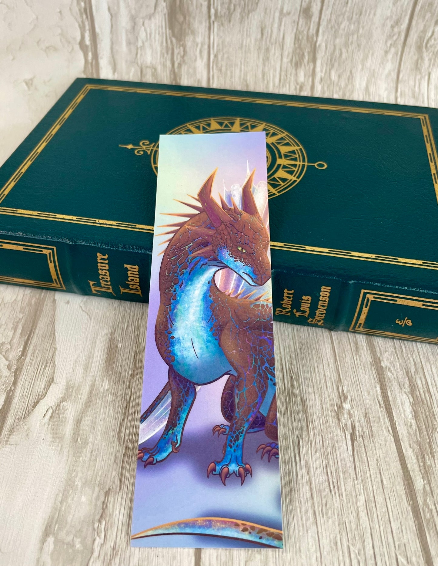 A 2 by 7 inch bookmark featuring a sitting, side profile of a dragon that has the features of a boulder opal with hues of blue, green, purple, and pink on the underbelly and cracks of the rough, brown hue, rock scales. The wings are tucked back and are of an iridescent shimmery hue that glitters in the light. The background is of a rainbow ombre effect