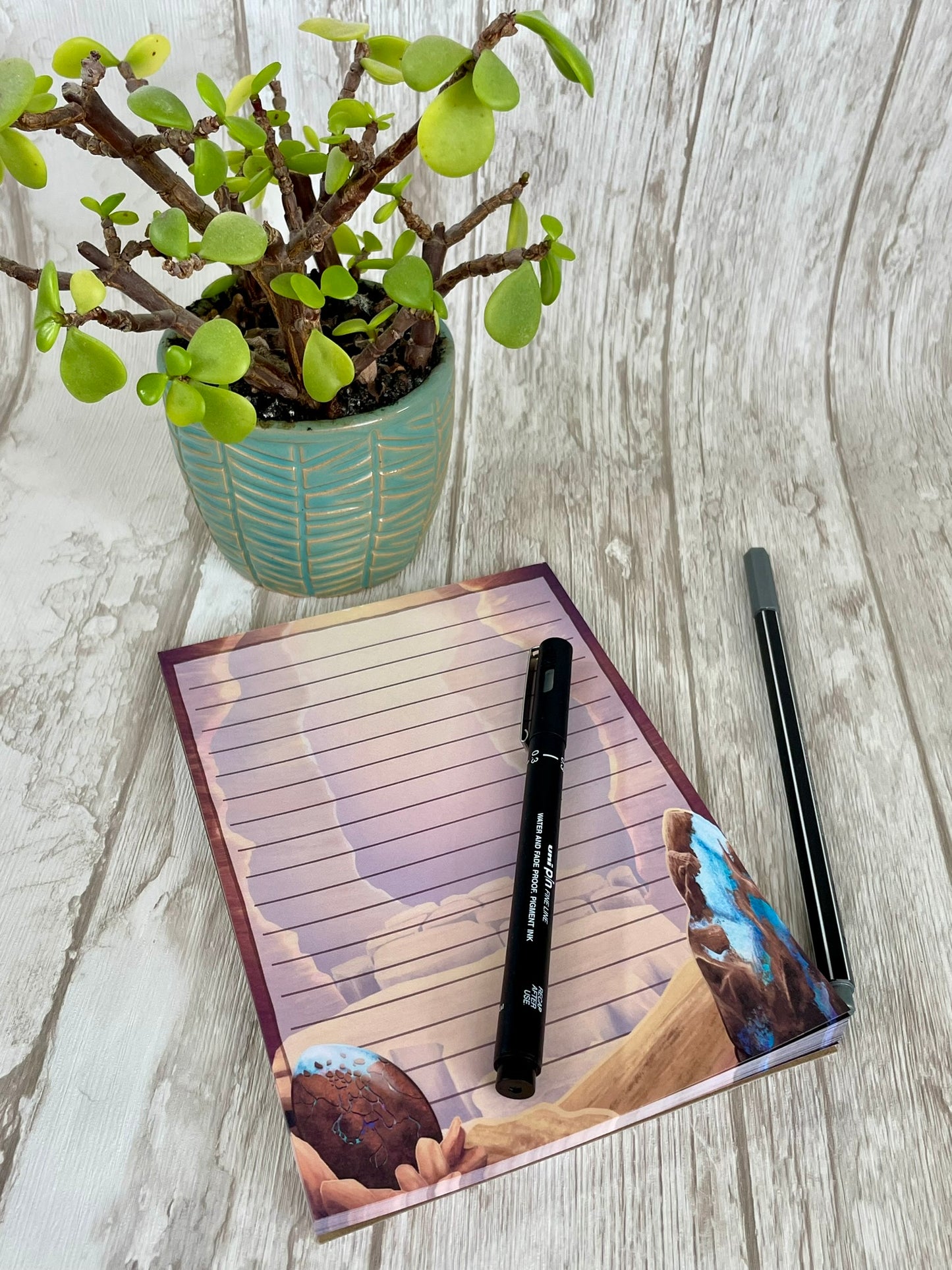 A 5 by 7 inch notepad with 50 pages that each feature the mesa canyon that is the background from the art print. Each page background has lines in which to write on and shows off the opal dragon egg in the left bottom hand corner and a large raw opal mineral rock on the bottom right hand corner.