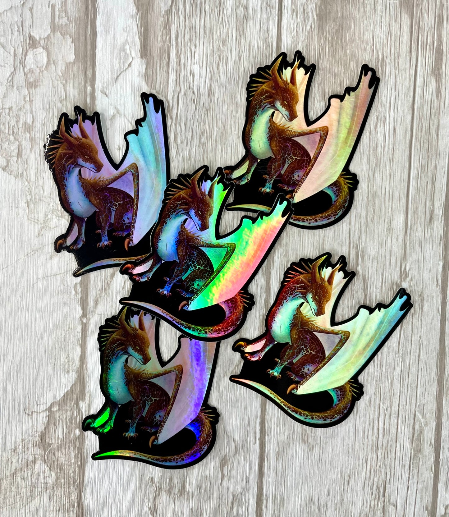 Multiple die cut, holographic, vinyl stickers with a black background featuring a sitting, side profile of a dragon that has the features of a boulder opal with hues of blue, green, purple, and pink on the underbelly and cracks of the rough, brown hue, rock scales. The wings are tucked back and are of an iridescent shimmery hue.