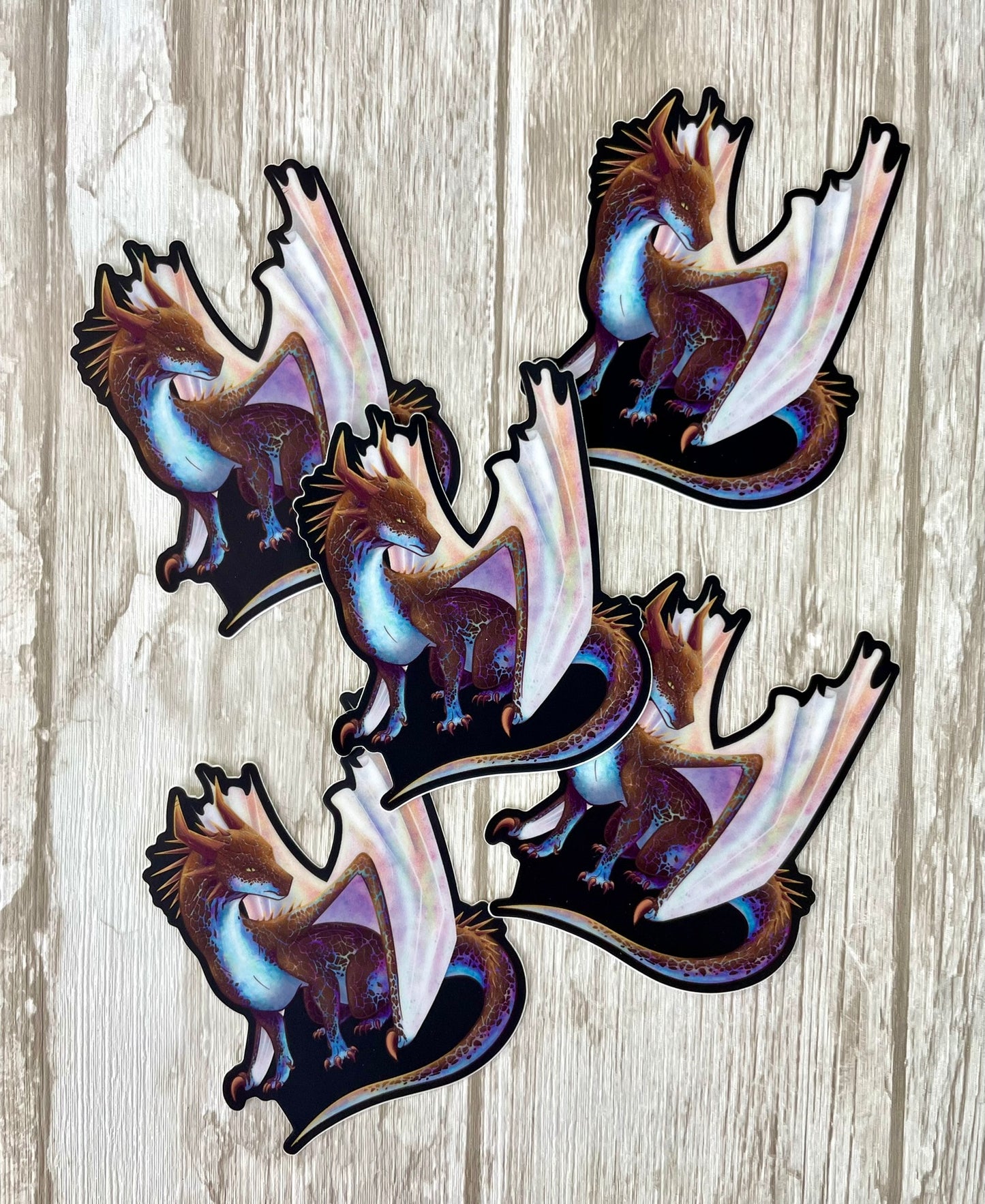 Multiple die cut, vinyl stickers with a black background featuring a sitting, side profile of a dragon that has the features of a boulder opal with hues of blue, green, purple, and pink on the underbelly and cracks of the rough, brown hue, rock scales. The wings are tucked back and are of an iridescent shimmery hue.