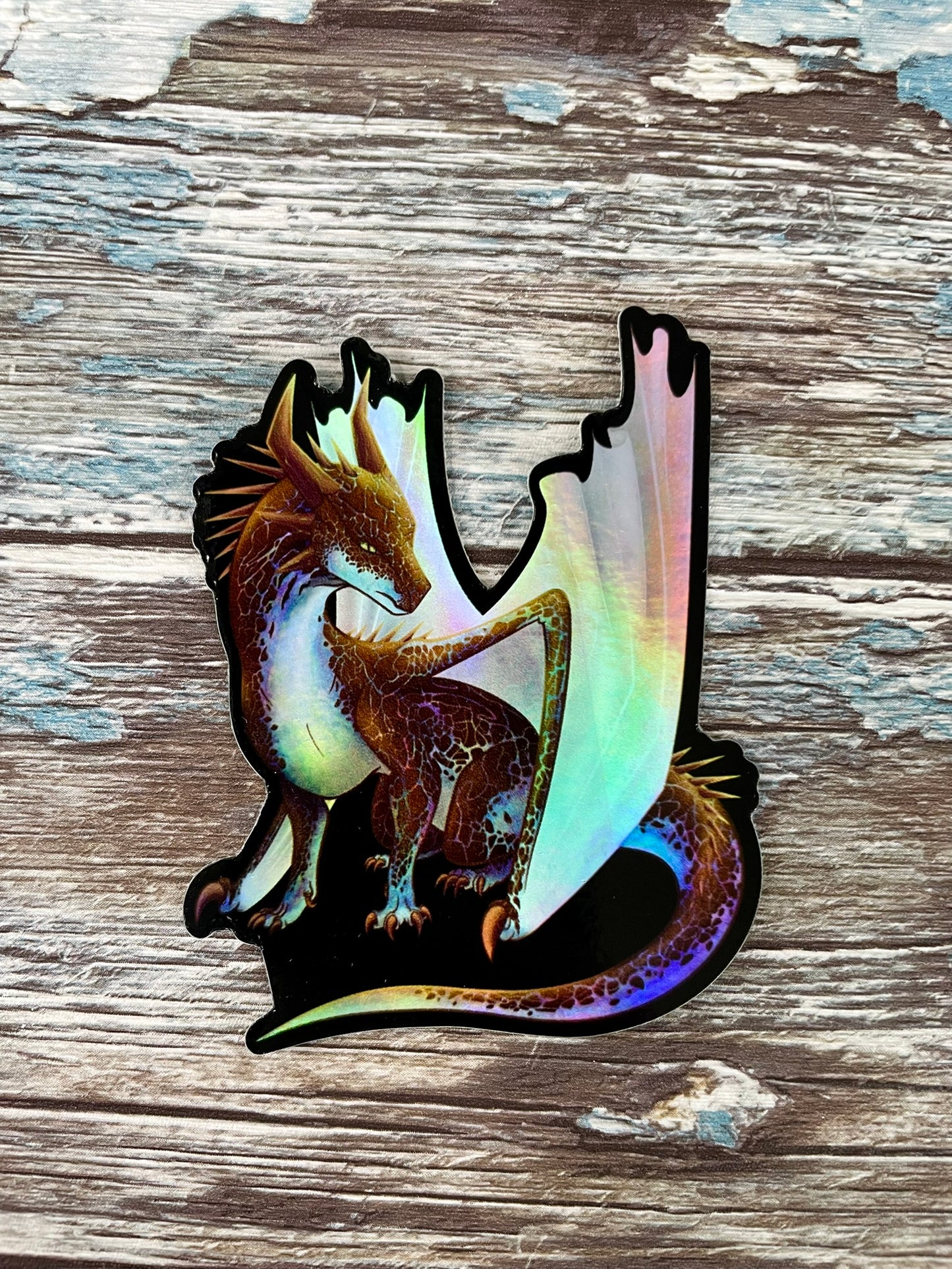 A die cut, holographic, vinyl sticker with a black background featuring a sitting, side profile of a dragon that has the features of a boulder opal with hues of blue, green, purple, and pink on the underbelly and cracks of the rough, brown hue, rock scales. The wings are tucked back and are of an iridescent shimmery hue.