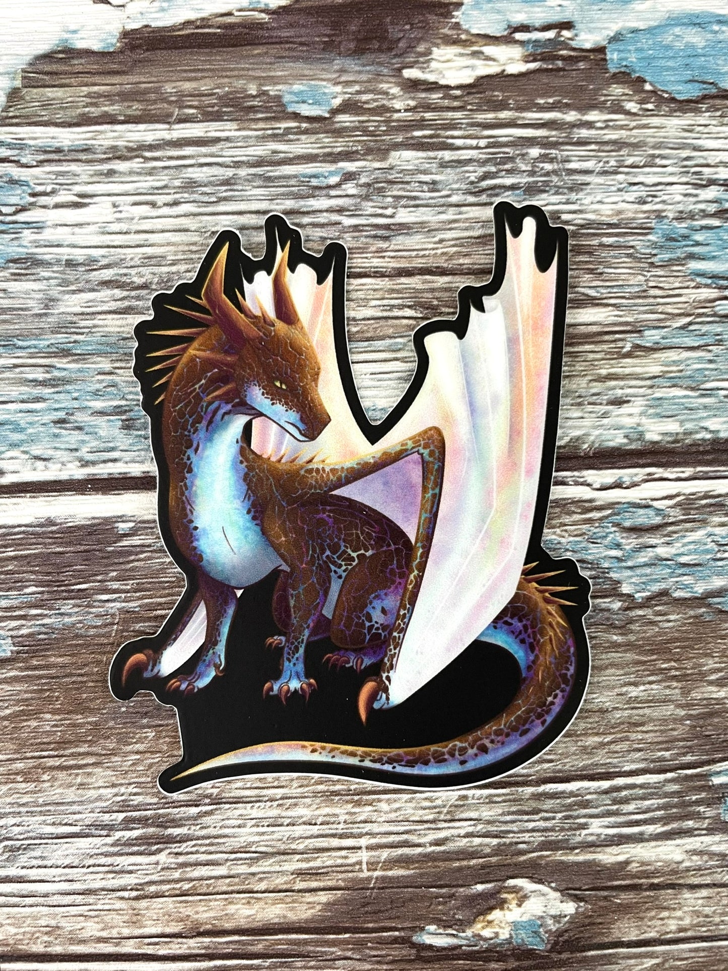 A die cut, vinyl sticker with a black background featuring a sitting, side profile of a dragon that has the features of a boulder opal with hues of blue, green, purple, and pink on the underbelly and cracks of the rough, brown hue, rock scales. The wings are tucked back and are of an iridescent shimmery hue.