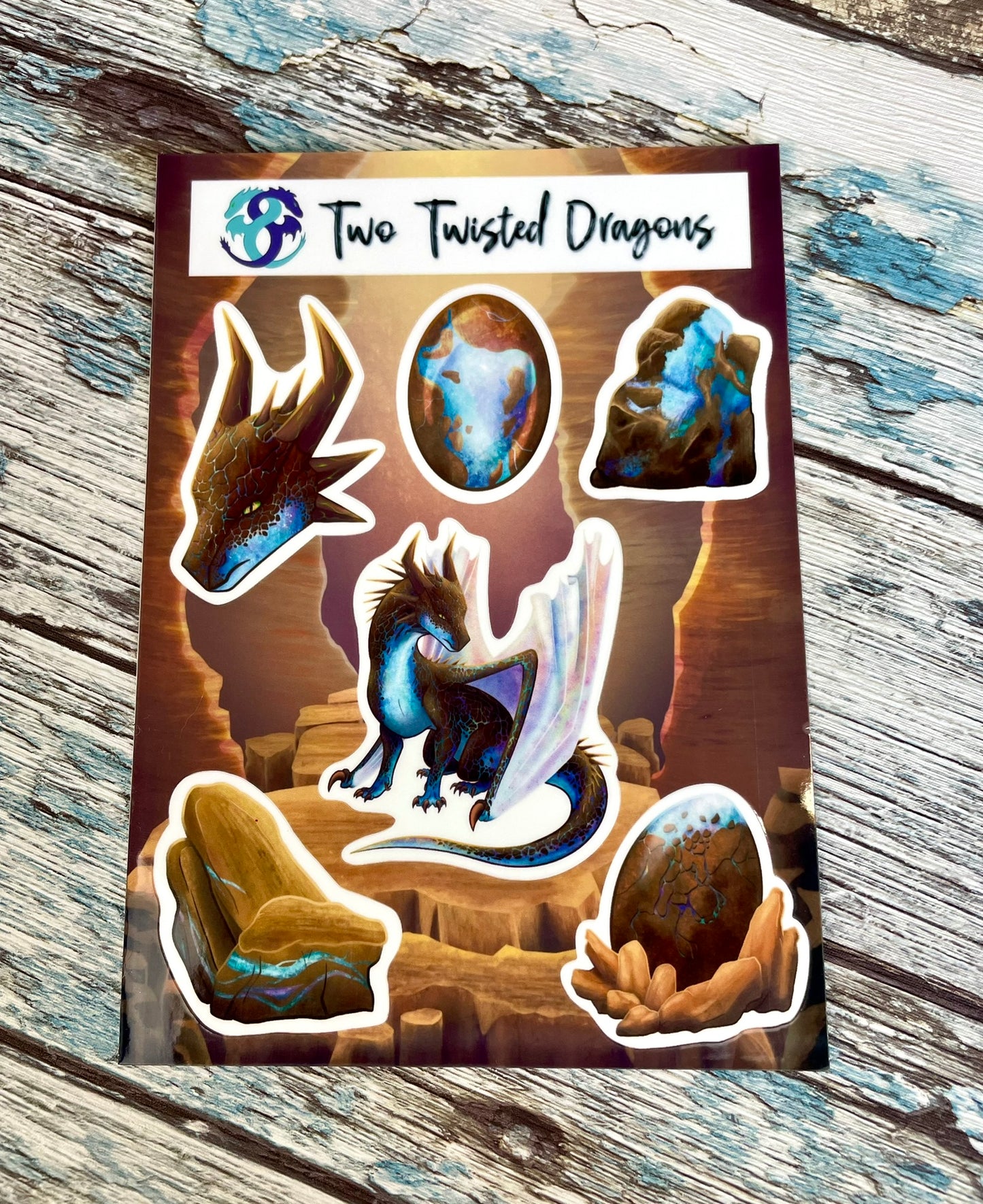 A 4x6 inch sticker sheet with 6 small different stickers featuring a sitting, side profile of a dragon that has the features of a boulder opal with hues of blue, green, purple, and pink on the underbelly and cracks of the rough, brown hue, rock scales. The wings are tucked back and are of an iridescent shimmery hue. There is also a head of the dragon, a dragon egg, two raw opal minerals, and one cut opal gem.