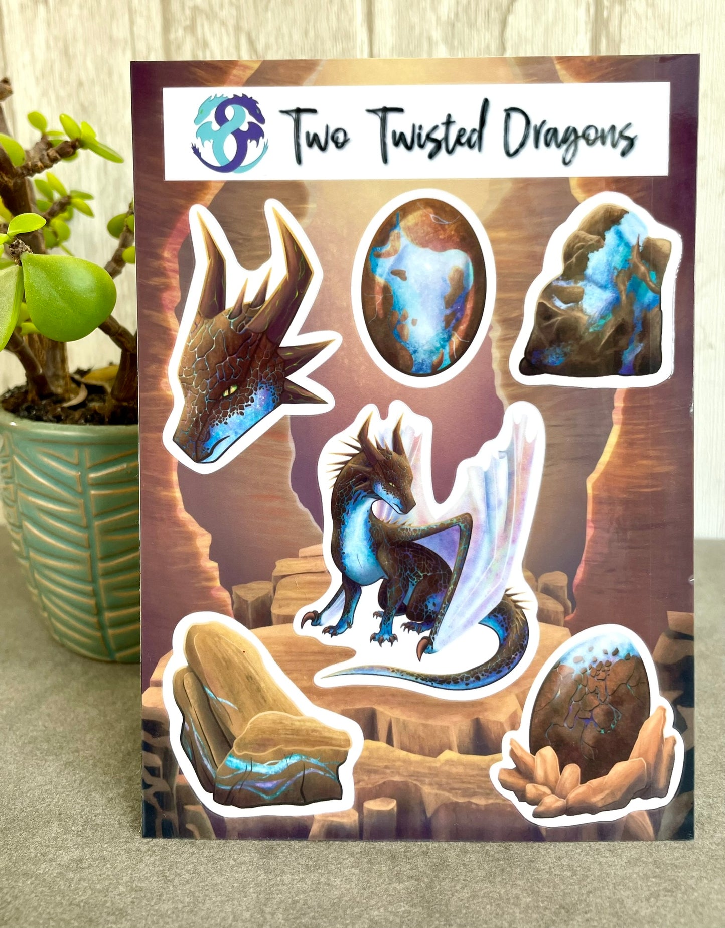 A 4x6 inch sticker sheet with 6 small different stickers featuring a sitting, side profile of a dragon that has the features of a boulder opal with hues of blue, green, purple, and pink on the underbelly and cracks of the rough, brown hue, rock scales. The wings are tucked back and are of an iridescent shimmery hue. There is also a head of the dragon, a dragon egg, two raw opal minerals, and one cut opal gem.