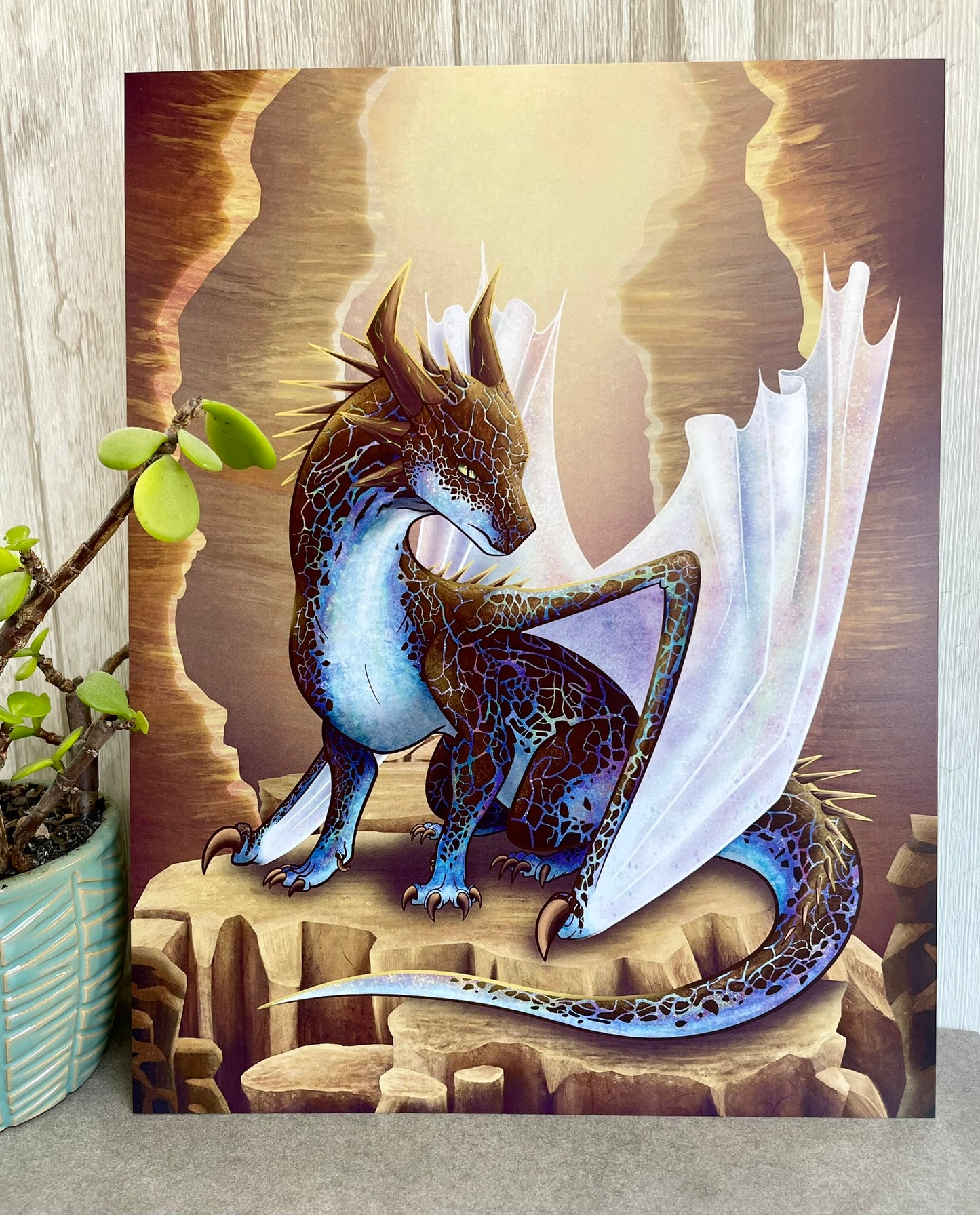 A 8 1/2 x 11 art print featuring a sitting, side profile of a dragon that has the features of a boulder opal with hues of blue, green, purple, and pink on the underbelly and cracks of the rough, brown hue, rock scales. The wings are tucked back and are of an iridescent shimmery hue. The background of the print is that of a mesa canyon with the dragon sitting on a large boulder within the canyon.