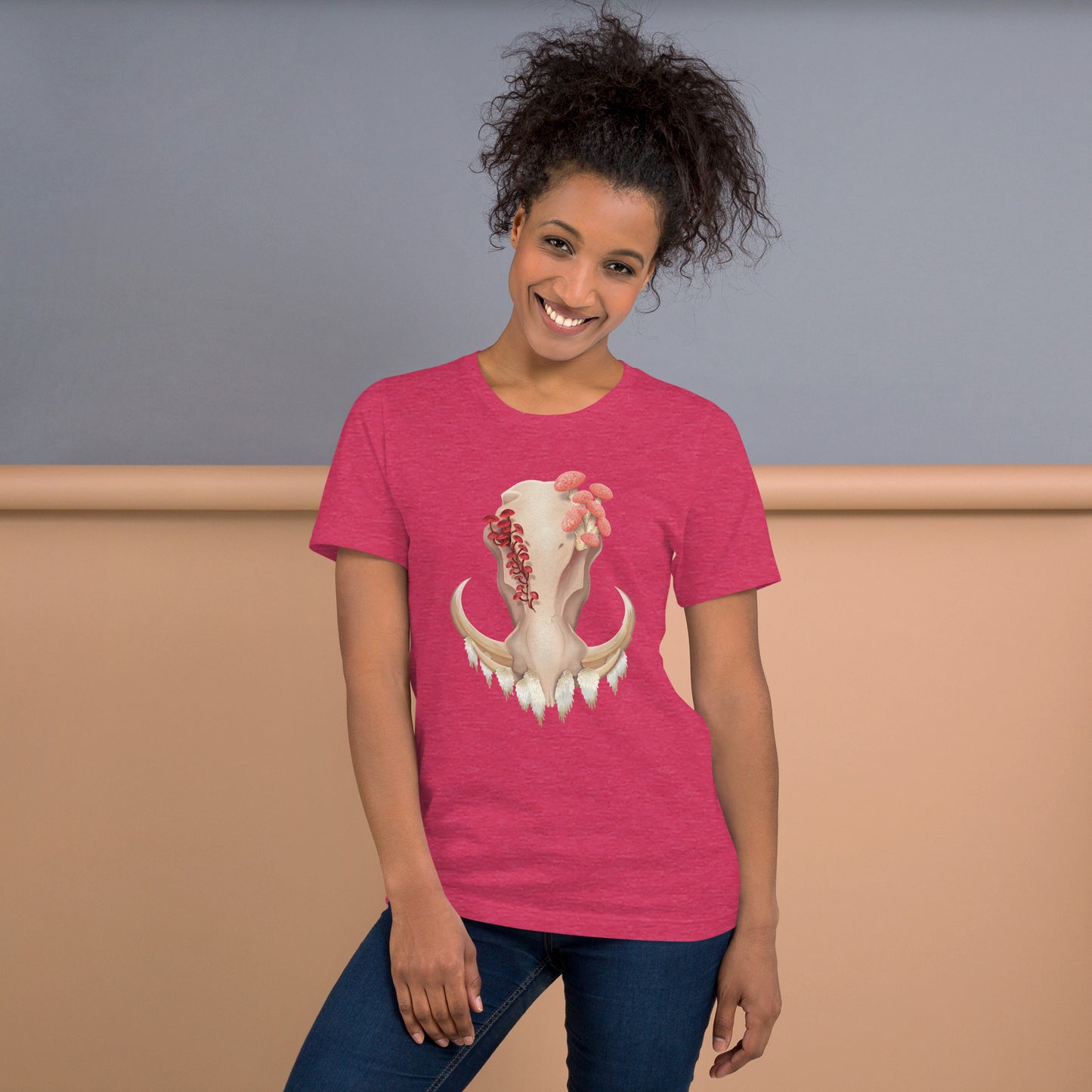 An adult wearing a heather raspberry colored, unisex T shirt featuring a front facing warthog skull with 3 different kinds of fungi growing out of it