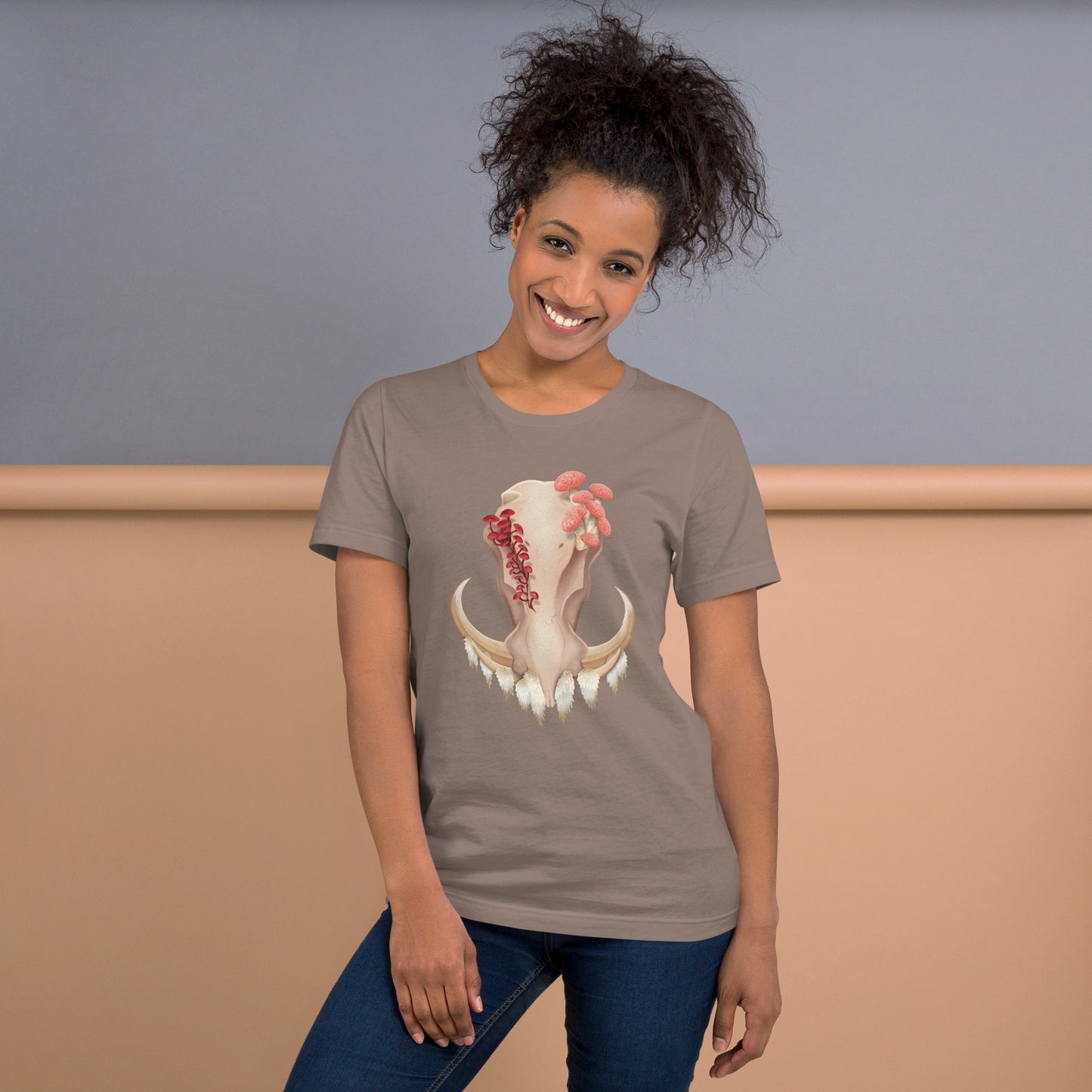 An adult wearing a pebble colored, unisex T shirt featuring a front facing warthog skull with 3 different kinds of fungi growing out of it