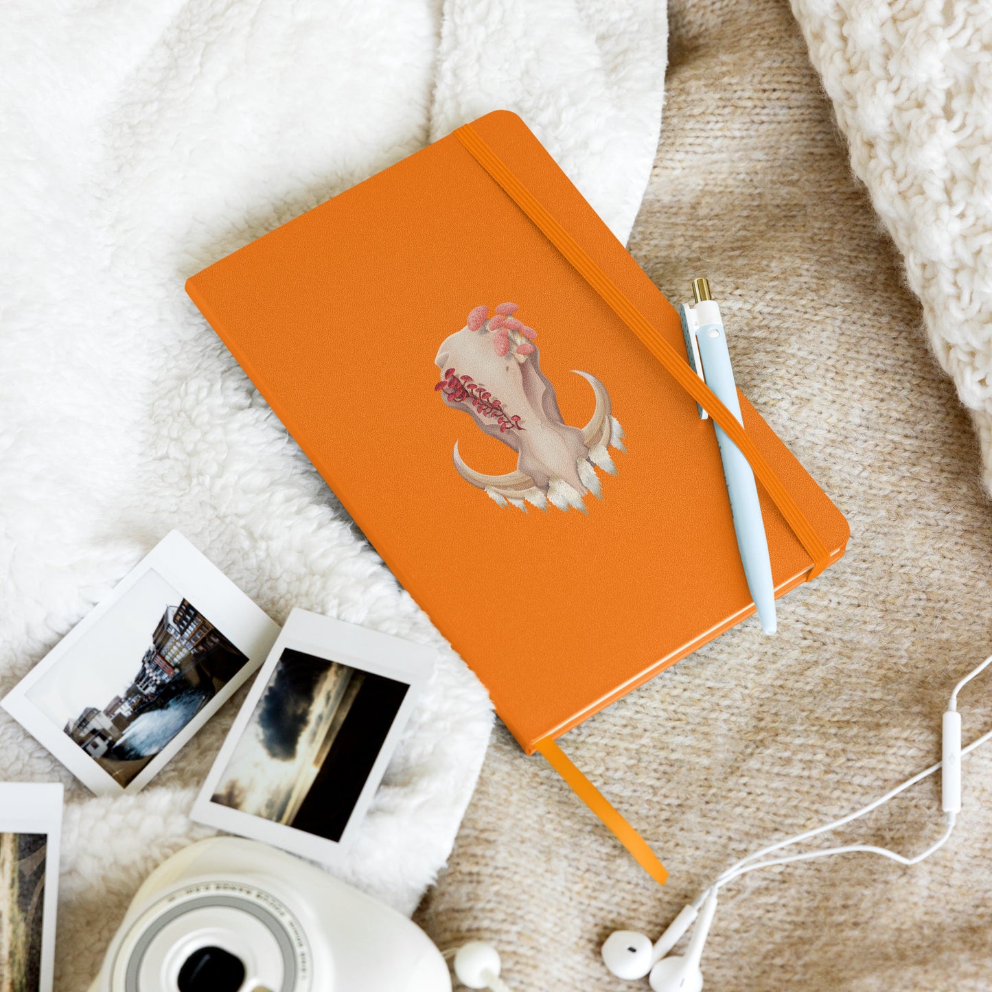 A 80 page, orange hardback lined journal with a pocket in the back, a ribbon book mark, and elastic closure, featuring a front facing warthog skull with 3 different kinds of fungi growing out of it
