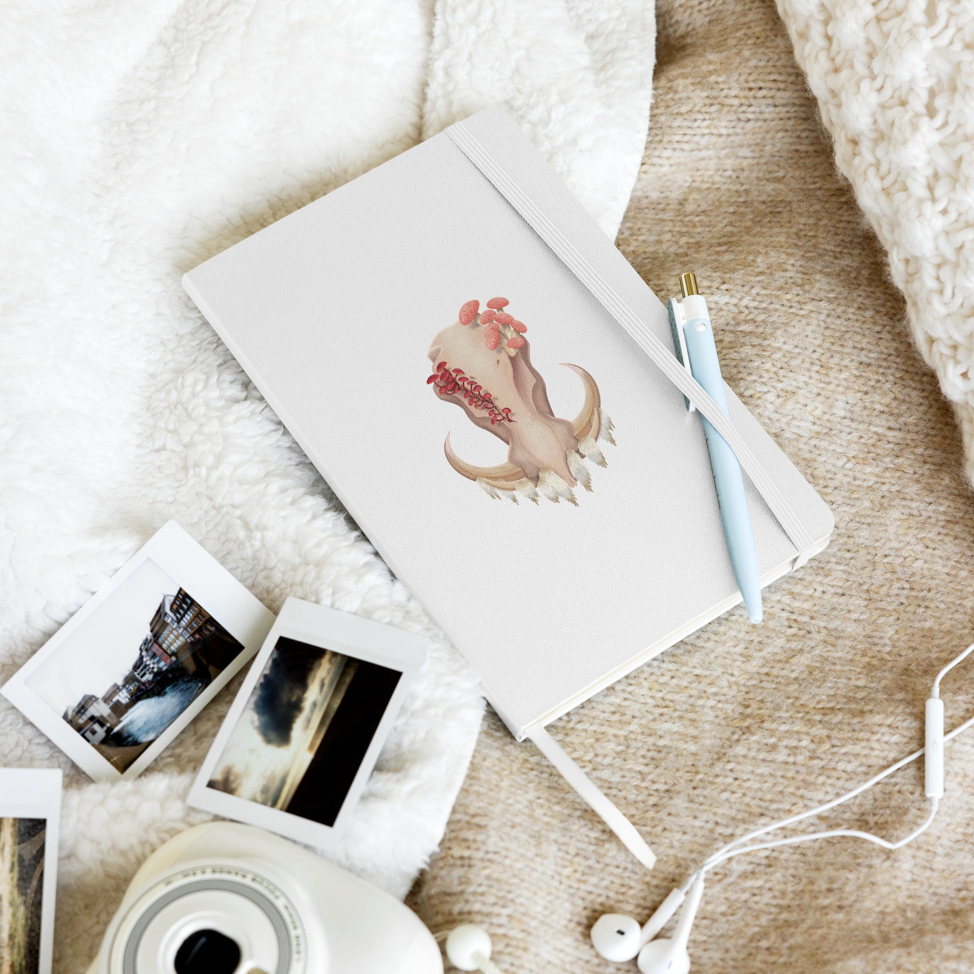 A 80 page, white hardback lined journal with a pocket in the back, a ribbon book mark, and elastic closure, featuring a front facing warthog skull with 3 different kinds of fungi growing out of it