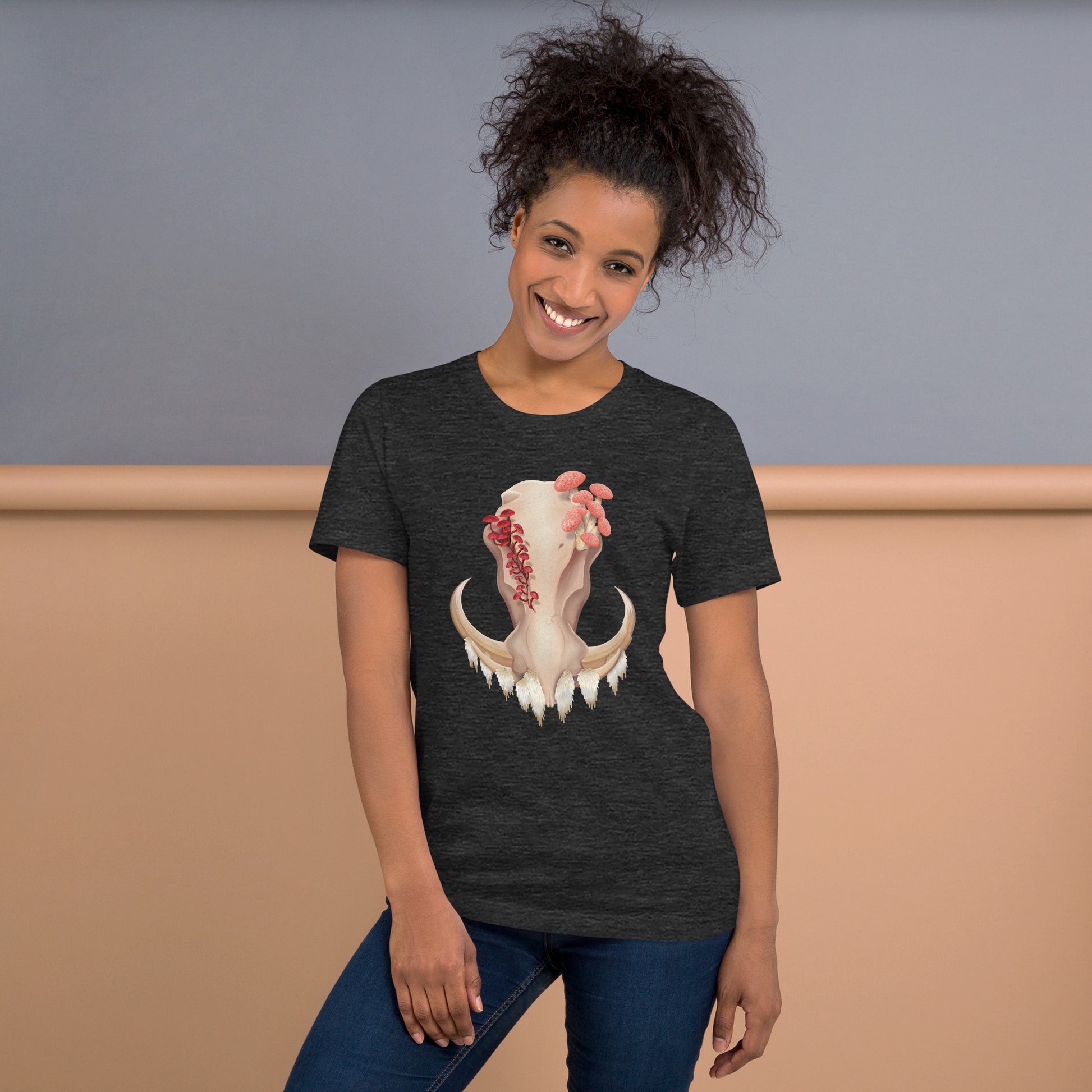 An adult wearing a dark heather grey colored, unisex T shirt featuring a front facing warthog skull with 3 different kinds of fungi growing out of it