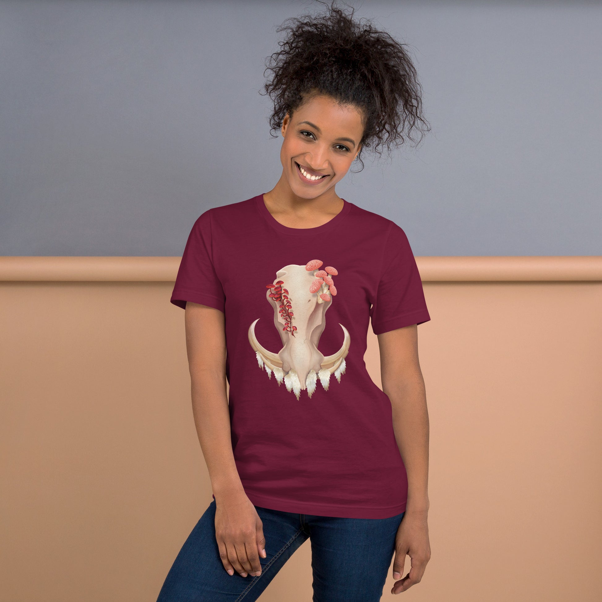 An adult wearing a maroon colored, unisex T shirt featuring a front facing warthog skull with 3 different kinds of fungi growing out of it