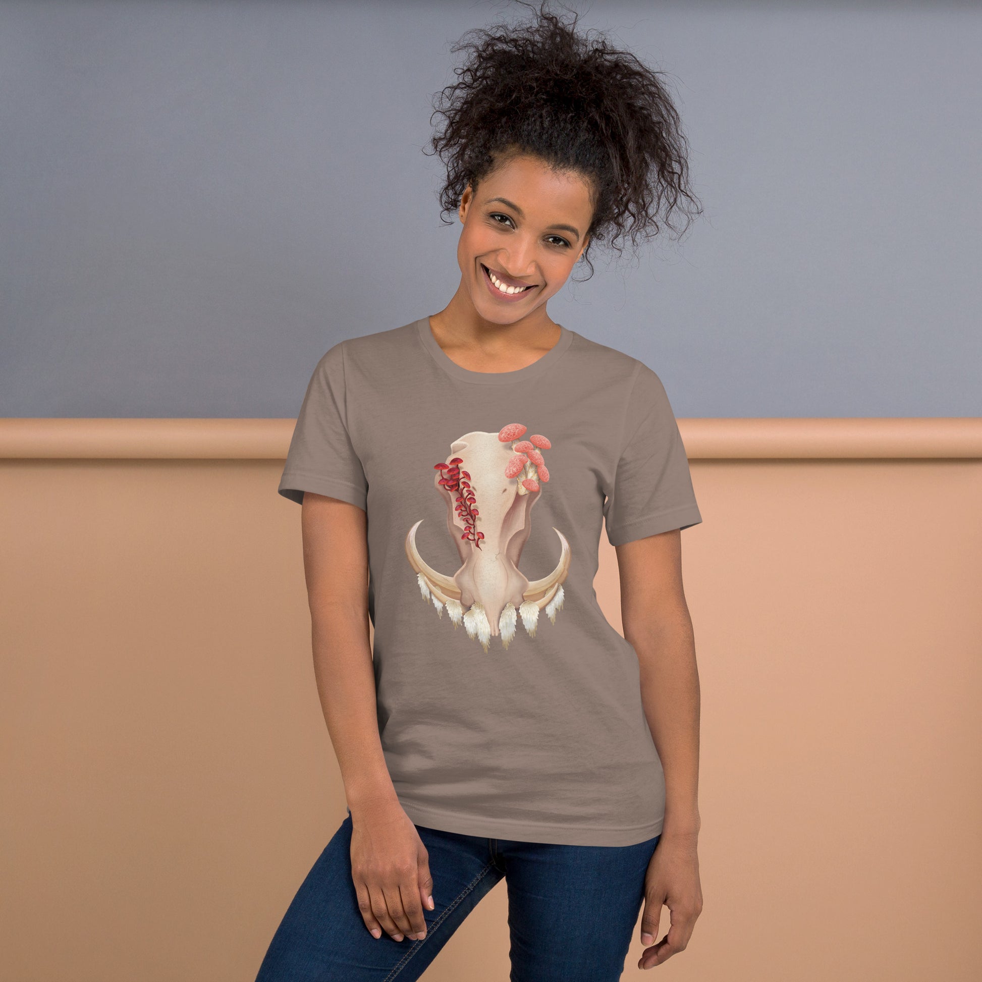 An adult wearing a pebble colored, unisex T shirt featuring a front facing warthog skull with 3 different kinds of fungi growing out of it