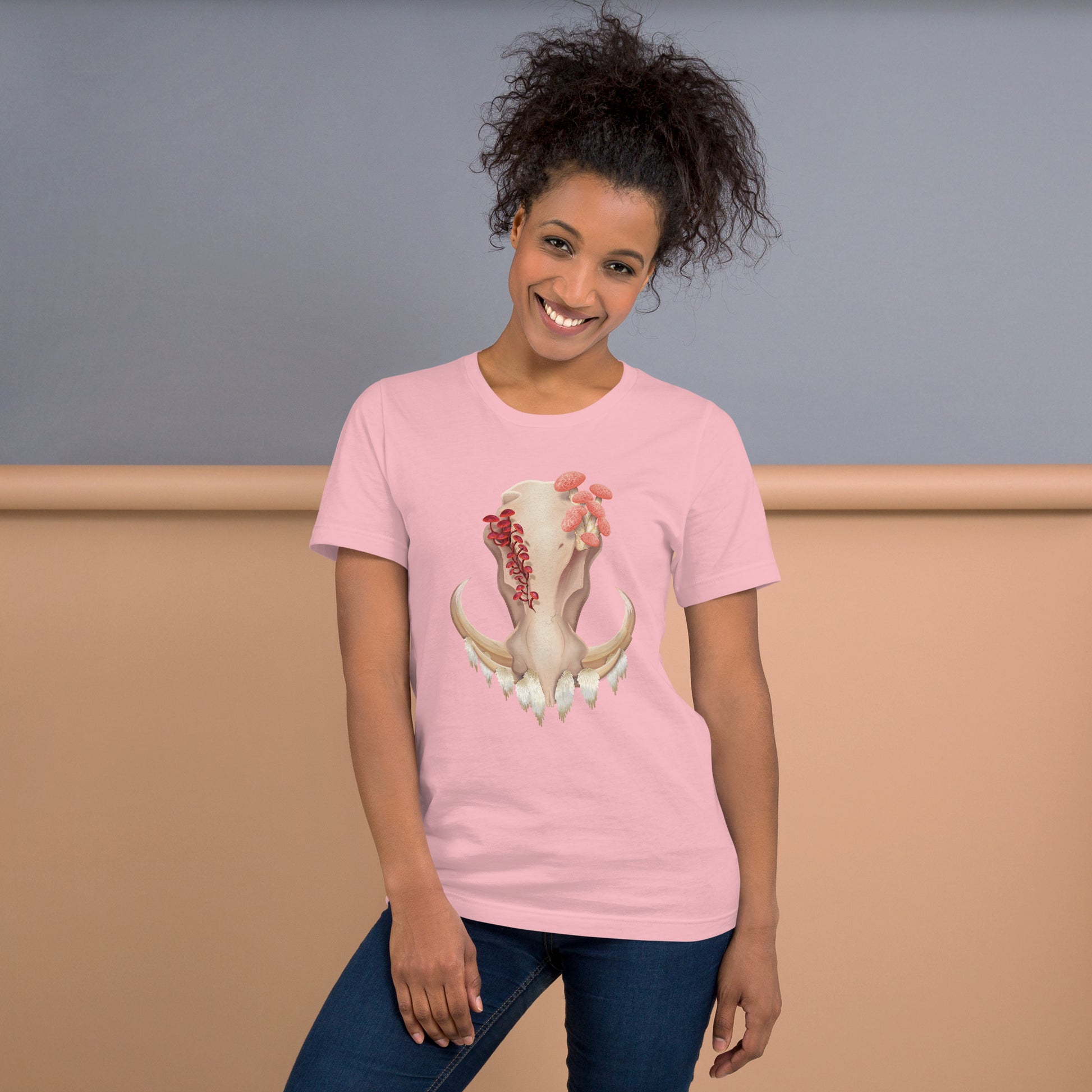 An adult wearing a light pink colored, unisex T shirt featuring a front facing warthog skull with 3 different kinds of fungi growing out of it