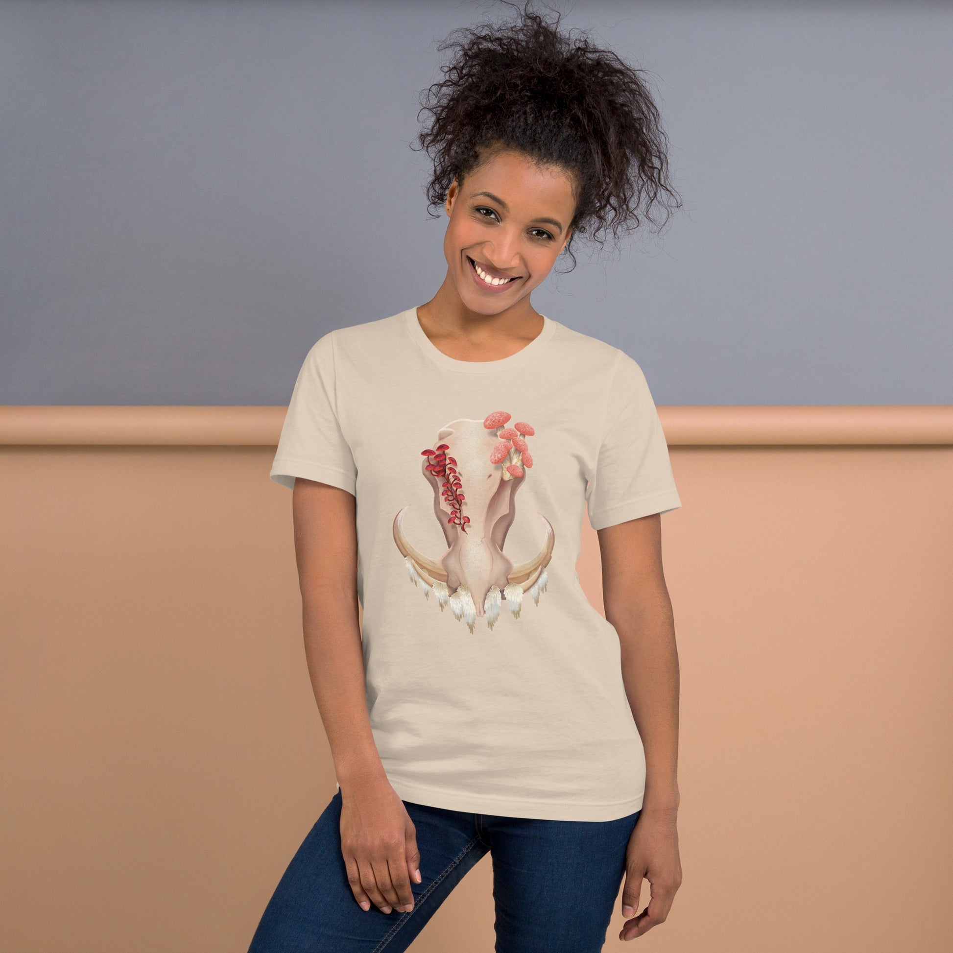 An adult wearing a soft cream colored, unisex T shirt featuring a front facing warthog skull with 3 different kinds of fungi growing out of it